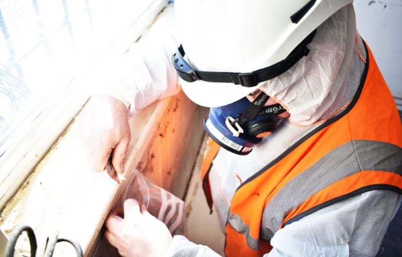Asbestos Testing Procedure Worker in protective gear conducting asbestos testing near a window with a sample collection tool.